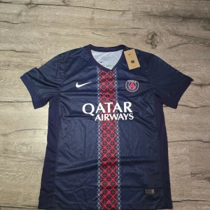 PSG fotbollströja - Paris Saint-Germain fotbollströja från Nike i marinblått med rött och vitt geometriskt mönster längs mitten. Kortärmad med V-ringad krage, PSG-logga på bröstet och Qatar Airways-tryck. Tillverkad i 100% polyester, perfekt för dig som älskar fotboll och vill ha en snygg supportertröja.