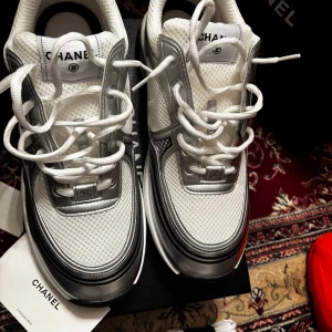 Chanel vita och silver sneakers - Snygga sneakers från Chanel i vitt och silver med mesh- och skinnpaneler. Klassisk Chanel-logga på sidan, tjock sula och snörning framtill. Perfekta för dig som vill ha en exklusiv och trendig look.