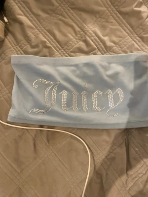 Ljusblå bandeau tubtopp Juicy Couture - Säljer en ljusblå bandeau tubtopp från Juicy Couture med glittrande strasstenar som stavar 'Juicy' framtill. Toppen är axelbandslös och har en mjuk, stretchig känsla som sitter snyggt på kroppen. Perfekt för dig som vill ha en ikonisk Y2K-look.