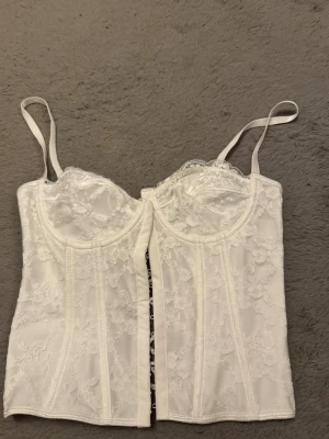 Vit spetsväst med snörning - Säljer en vit väst med tunna axelband och vacker spetsdetalj. Västen har snörning i sidan och hakar framtill för stängning. Sitter tajt och har en feminin, elegant look med fina spetsmönster över hela plagget.