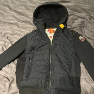 Svart Parajumpers pufferjacka S - Svart pufferjacka från Parajumpers med huva och broderad patch på ärmen. Jackan har dragkedja framtill, två sidofickor med dragkedja och detaljer i orange. Materialmix av fleece och puffer ger både värme och stil. Perfekt för kyliga dagar. Köpt på johnells för 4000. 