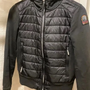 Svart pufferjacka från Parajumpers - Svart pufferjacka från Parajumpers med quiltad front och släta ärmar. Jackan har dragkedja framtill, två sidofickor med dragkedja och en snygg patch på ena ärmen. Perfekt för dig som gillar stilren och sportig look.