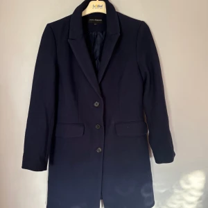 Mörkblå kappa från Kappahl - Snygg mörkblå kappa från Kappahl med klassisk krage och tre knappar framtill. Jackan har två fickor med lock och är helfodrad. Materialet är en mix av polyester och viskos med lite elastan för extra komfort. Perfekt för dig som gillar stilrena och tidlösa plagg.