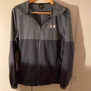 Vindjacka Under Armour - Snygg tvåfärgad vindjacka från Under Armour i mörkgrå och ljusgrå toner. Jackan har huva, dragkedja framtill och en vit logga på bröstet. Perfekt för dig som gillar sportig stil och vill ha något lätt och funktionellt. Justerbar huva och diskreta detaljer.