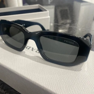 Svarta solglasögon från Prada - Snygga svarta solglasögon från Prada med rektangulära bågar och breda skalmar. Glasögonen har en modern och edgy look med diskret logga på sidan. Perfekta för dig som vill sticka ut med en stilren accessoar.