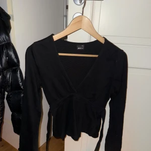 Svart omlottopp från Gina Tricot XS - Svart omlottopp från Gina Tricot i storlek XS. Toppen har lång ärm, v-ringad hals och knytband i midjan för en snygg passform. Materialet är mjukt och stretchigt, perfekt för att skapa en cool och stilren look.