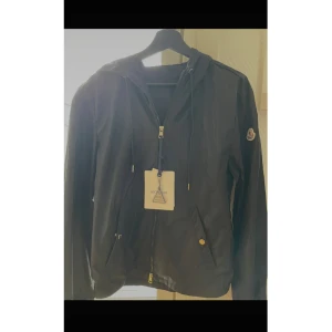 Moncler Windbreaker  - Svart vindjacka från Moncler med huva och snörning, dragkedja framtill och två fickor med tryckknappar. Jackan har Moncler-logga på ärmen och diskret rödvit detalj på huvan. Materialet är lätt och syntetiskt, perfekt för blåsiga dagar. Helt oanvänd. Storlek 2