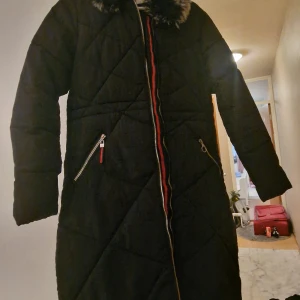 Svart lång dunjacka med fuskpäls - Snygg svart lång dunjacka från A.X.J.W med quiltad design och fuskpäls vid kragen. Jackan har två snedställda fickor med dragkedja och en färgglad detalj längs dragkedjan framtill. Perfekt för kalla dagar och har en stilren look.