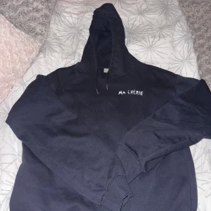 Svart hoodie Ma Chérie Gina Tricot - Svart hoodie från Gina Tricot med vit text 'MA CHÉRIE' och hjärtmotiv på ryggen. Tröjan har huva med snörning och långa ärmar. Mjuk insida i bomullsmix, perfekt för chill dagar. Snygg och enkel att matcha med jeans eller leggings.