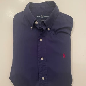 Mörkblå skjorta från Ralph Lauren - Klassisk mörkblå skjorta från Ralph Lauren med röd broderad logga på bröstet. Skjortan har lång ärm, knappar hela vägen och button-down krage. Classic fit-modell i mjuk bomull, perfekt för en clean och stilren look. Jag har ett erbjudande om man köper min andra Ralph lauren skjorta också så får man båda dem för 499, istället för 699🙌