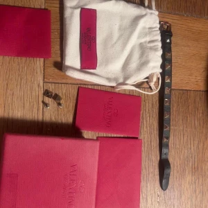 Valentino svart armband med nitar - Snyggt svart armband från Valentino med guldiga pyramidnitar. Armbandet är smalt och har en cool rockig vibe, perfekt för att lyfta din outfit. Kommer med originalförpackning och dustbag från Valentino.