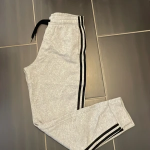 Grå mjukisbyxor från Adidas - Snygga grå mjukisbyxor från Adidas med svarta ränder längs sidorna och svart snörning i midjan. Byxorna har elastisk midja och är tillverkade i mjukt bomullsmaterial, perfekta för chill eller träning.