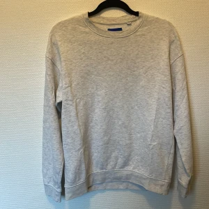Ljusgrå sweatshirt från Jack & Jones - En stilren ljusgrå sweatshirt från Jack & Jones Originals. Tröjan har rund hals, ribbade muddar vid ärmslut och nederkant samt långärmad passform. Perfekt för chill dagar och enkel att matcha med jeans eller joggers.