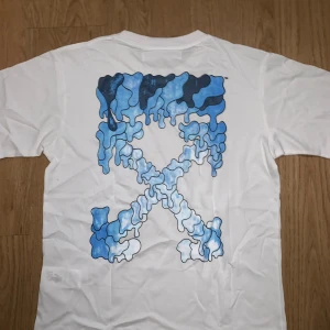 Off-White vit t-shirt med blått tryck - Säljer en vit Off-White t-shirt med korta ärmar och rund hals. Framsidan har svart Off-White-logga med handmotiv, medan baksidan har ett stort blått och mörkblått grafiskt tryck i Off-Whites klassiska X-form. Materialet är mjuk bomull, perfekt för dig som gillar streetwear.