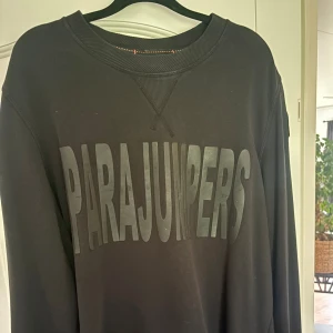 Svart sweatshirt från Parajumpers - Svart sweatshirt från Parajumpers med stort tryck över bröstet och patch på ärmen. Tröjan har rund hals, ribbade muddar och långa ärmar. Materialet är mjuk bomull och passformen är normal. Perfekt för dig som gillar stilrena och sportiga plagg.