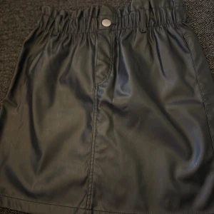 Svart kort kjol i skinnimitation - Säljer en svart kort kjol i skinnimitation med snygg rynkad midja och knapp framtill. Kjolen har en clean look och passar perfekt till sneakers eller boots. Den är enkel att matcha och har en cool vibe som lyfter din outfit. Är storlek 152 men passar som XS.