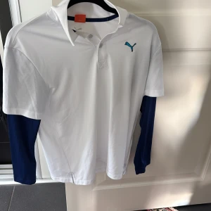 Vit pikétröja från Puma med blå detaljer - Snygg vit pikétröja från Puma med klassisk krage och knappar framtill. Tröjan har blå långa ärmar under den kortärmade pikén, samt Puma-logga på bröstet och diskreta svarta prickar på axeln. Perfekt för sportiga tillfällen eller en chill look.
