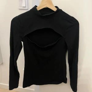 Svart långärmad polotopp med öppning - Cool svart långärmad polotopp med unik öppen detalj över bröstet. Toppens material känns mjukt och stretchigt, perfekt för dig som gillar statement-plagg med edge. Passformen är slim och toppen har en hög krage som ger en snygg siluett.