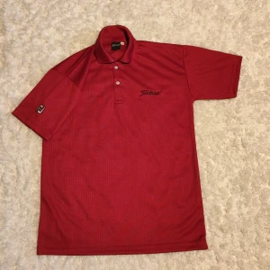 Röd Titleist piké med diskret mönster - Röd pikétröja från Titleist med subtilt randigt mönster, klassisk krage och två knappar framtill. Broderad Titleist-logga på bröstet och liten logga på ärmen. Perfekt för golf eller chill dagar. Tillverkad i mjukt och lätt material.