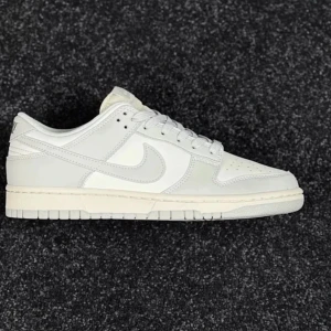 Nike Dunk Low vit/grå sneakers - Nike Dunk Low sneakers i en clean vit och ljusgrå färg. Klassisk siluett med rund tå, platt sula och snörning. Swoosh-loggan på sidan och paneler i skinn ger en stilren vibe. Perfekt för dig som gillar minimalistisk streetstyle och vill ha ett par fräscha kicks.