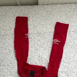 Röda Umbro fotbollsstrumpor - Säljer ett par röda fotbollsstrumpor från Umbro med vit logga på sidan. Strumporna är långa och har en klassisk sportig look, perfekta för match eller träning. 