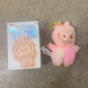 Pop Mart Big Into Energy Tiny Plush Pendant - Söt och samlarvänlig Pop Mart Big Into Energy Tiny Plush Pendant. Mjuk, fluffig figur i rosa och vitt med nyckelringsfäste. Levereras med originalförpackning, ser ut att vara i nyskick utan synligt slitage. Perfekt för samlare eller som present.