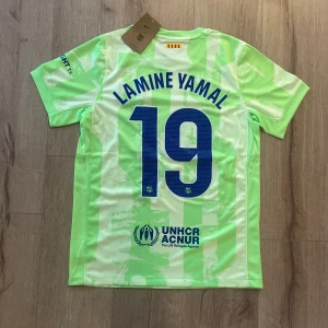 Barcelona Lamine Yamal fotbollströja - Säljer en officiell FC Barcelona fotbollströja med Lamine Yamal och nummer 19 på ryggen. Tröjan är ljusgrön med vita inslag i ett unikt mönster, har korta ärmar och är tillverkad i lätt polyester. Nike-logga, Spotify och UNHCR tryck samt klubbmärke på bröstet.