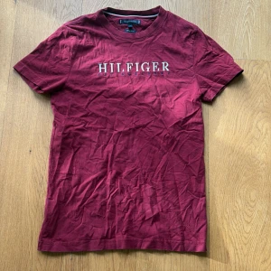 Vinröd Tommy Hilfiger t-shirt - Snygg vinröd t-shirt från Tommy Hilfiger i ekologisk bomull. Framsidan har broderad Hilfiger-logga i vitt och blått. Klassisk rund hals och korta ärmar. Perfekt för dig som gillar stilrena märkesplagg med tydlig branding.