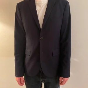 Mörkblå kavaj  - Snygg mörkblå kavaj med enkelknäppning och klassisk krage. Modellen har två fickor med lock framtill och en bröstficka. Passformen är normal och tyget känns mjukt och stilrent. Perfekt för dig som vill ha en clean och tidlös look.