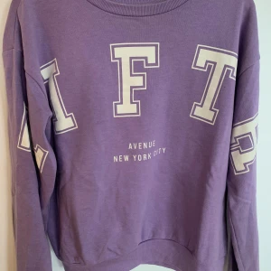 Lila sweatshirt med tryck NYC - Lila sweatshirt med stora vita bokstäver och texten 'Avenue New York City' framtill. Tröjan har rund hals, ribbade muddar och lång ärm. Perfekt för en chill och avslappnad stil.