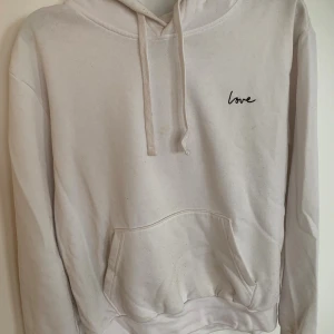Vit hoodie med broderad text Love - Vit hoodie med broderad svart text 'Love' på bröstet. Tröjan har huva med snörning och en stor magficka framtill. Tillverkad i mjuk bomull och har långa ärmar. Perfekt för en chill och avslappnad stil.