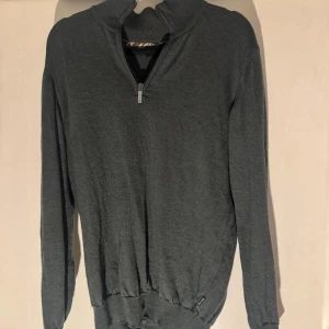Mörkgrön stickad half zip från Sand - Snygg mörkgrön stickad tröja från Sand Copenhagen med half zip och långärmad design. Tröjan har en ribbad kant nedtill och vid ärmslut, samt en cool detalj med mönstrad insida vid kragen. Perfekt för dig som gillar stilrena och bekväma plagg.