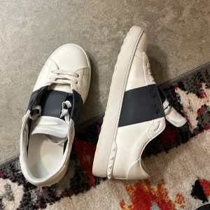 Valentino Garavani vita sneakers - Snygga vita Valentino Garavani sneakers med bred svart rem över vristen och coola nitar på hälen. Skorna har rund tå, snörning och är gjorda i skinn. Perfekta för dig som vill ha en stilren men ändå lyxig look.