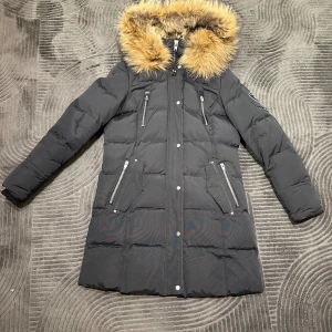 Svart dunjacka med päls rockandblue - Snygg svart lång dunjacka från Rock and blue  med fluffig päls  på huvan. Jackan har flera dragkedjefickor,  och långa ärmar. Perfekt för kalla dagar och har en stilren look med silverdetaljer. Den är använd endast 1 vinter 