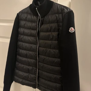 Svart  cardigan från Moncler XS - Svart ä cardigan från Moncler med quiltad glansig front och rygg i syntetmaterial, samt stickade ärmar och krage. Jackan har dragkedja framtill och Moncler-logga på ärmen. Perfekt för dig som vill ha en stilren och sportig look. Knappast använf nästan som ny därav inga defekter eller dofter. Slutsåld på monclers hemsida och kostar 9500 kr på ansra hemsidor. Mitt pris 5000 kr men kan diskuteras vid snabbköp