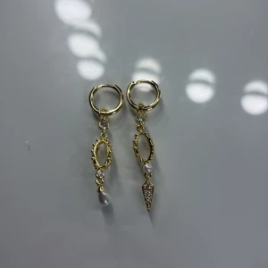 Guldiga örhängen med pärlor och stenar - Säljer ett par unika guldiga örhängen med hängande detaljer. Ena örhänget har en vit pärla och det andra har en spetsig hängande detalj med små glittriga stenar. 