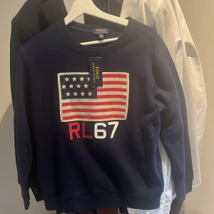Mörkblå sweatshirt Polo Ralph Lauren - Legat i min garderob ett tag men Aldrig använd, mörkblå sweatshirt från Polo Ralph Lauren med stort amerikanskt flaggtryck och RL67 framtill. Klassisk rund hals och långa ärmar. Skön och stilren tröja i bomull, perfekt för dig som gillar sportig och preppy stil.