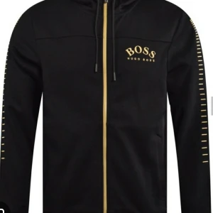 Svart hoodie från Hugo Boss L - Snygg hoodie från Hugo Boss med hel dragkedja och huva. Använd 2-3 gånger. Tröjan har gulddetaljer längs ärmarna och guldloggan på bröstet. Tillverkad i mjuk bomull och har ribbade muddar. Perfekt för en chill och stilren look.