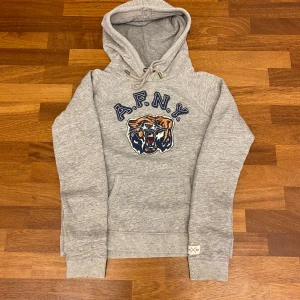 Grå hoodie Abercrombie & Fitch tiger - Snygg grå hoodie från Abercrombie & Fitch 1892 Collection med broderad tiger och A.F.N.Y. på bröstet. Tröjan har huva med snörning och känguruficka framtill. Perfekt för chill dagar och streetstyle. En vintage och väldigt efterfrågad hoodie som passar som ett litet S