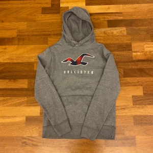 Grå hoodie från Hollister med logga - Säljer en grå hoodie från Hollister med stor logga och röd-rutigt mönster på bröstet. Tröjan har huva med snörning och en stor magficka. Perfekt för dig som gillar en avslappnad och snygg streetstil. Vintage och väldigt efterfrågat plagg.