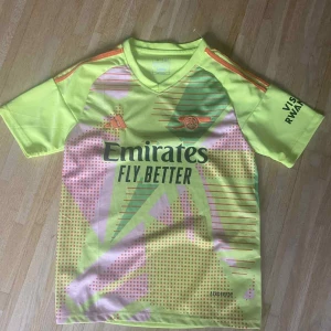Arsenal Adidas neon fotbollströja - Säljer en Arsenal fotbollströja från Adidas i neon-gul med rosa och gröna detaljer. Tröjan har korta ärmar, V-ringning och tryck med Emirates Fly Better på bröstet. Klassiska Adidas-ränder i orange på axlarna. Tillverkad i lätt och ventilerande polyester.