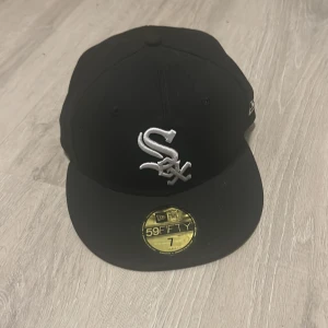 Svart New Era 59FIFTY White Sox keps - Säljer min svarta New Era 59FIFTY keps med Chicago White Sox-logga broderad i vitt framtill. Klassisk flat brim och MLB-logga bak. Kepsen är i modellen fitted och har detaljer i silver. Perfekt för dig som gillar streetwear och sportig stil. Den är helt oanvänd och i sprillans nytt skick, bara testad en gång då den var alldeles för liten. Storleken är 7 det vill säga  55.8cm.