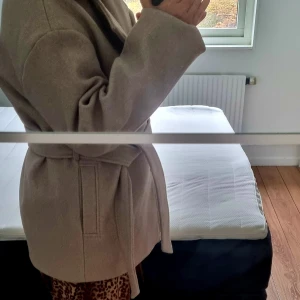 Beige kappa från NA-KD, stl 36 - Stilren beige kappa från NA-KD i storlek 36. Kappan har klassisk krage, knäppning framtill med knappar och ett bälte i midjan för snygg passform. Perfekt för kyligare dagar och enkel att matcha med olika outfits.