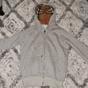 Grå Burberry hoodie - Säljer en grå hoodie från Burberry med klassiskt rutigt foder i huvan och broderad logga på bröstet. Tröjan har snörning i huvan, två fickor fram och dragkedja. Perfekt för dig som gillar stilrena och lyxiga detaljer.