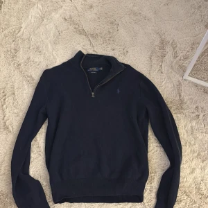 Marinblå half zip tröja Polo Ralph Lauren - Snygg marinblå tröja från Polo Ralph Lauren med half zip och klassisk krage. Tröjan har långärmad passform och är tillverkad i mjuk bomull. Diskret broderad logga på bröstet och ribbade muddar vid ärmslut och nederkant. Perfekt för en chill och stilren look.