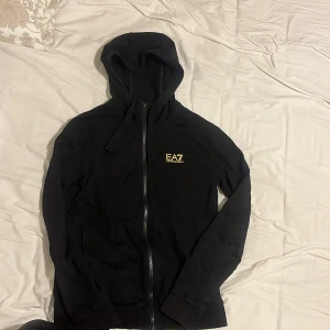 Svart hoodiejacka från EA7 Emporio Armani - Svart hoodiejacka från EA7 Emporio Armani med gul logotyp på bröstet. Jackan har dragkedja framtill, huva med snören och ribbade muddar vid ärmslut och nederkant. Perfekt för en sportig och stilren look.