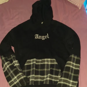 Svart hoodie - Svart hoodie från SHEIN med vit text 'Angel' på bröstet. Ärmar och magficka har svartvitt rutigt tyg som kontrast. Klassisk huva och lång ärm, perfekt för en cool streetstyle-look.