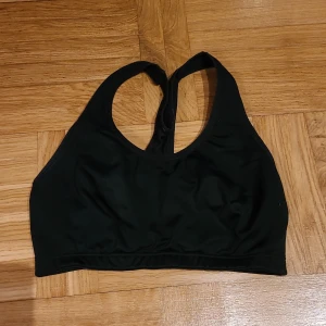 Svart sportig linnetopp - Säljer en svart sportig linnetopp med brottarrygg och elastisk nederkant. Toppen är tillverkad i ett stretchigt syntetmaterial som andas, perfekt för träning. Enkel och stilren design utan mönster eller extra detaljer.