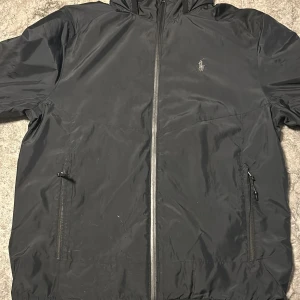 Svart vindjacka från Polo Ralph Lauren - Svart vindjacka från Polo Ralph Lauren med huva och dragkedja framtill. Jackan har två fickor med dragkedja och meshfoder på insidan. Klassisk logga broderad på bröstet och stilren design som funkar året runt. Perfekt för lite regniga dagar och blåsiga. Jackan är lite smutsig men inga större defekter. Skriv vid minsa fråga eller fundering. Priset är inte hugget i sten kan diskuteras vid snabb affär.