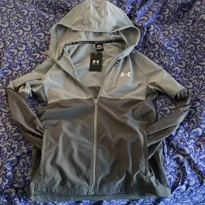Grå vindjacka från Under Armour - Snygg grå vindjacka från Under Armour med huva och dragkedja framtill. Jackan har en stilren design med logga på bröstet och är perfekt för aktiva dagar. Tillverkad i lätt polyester som står emot vind och väta.